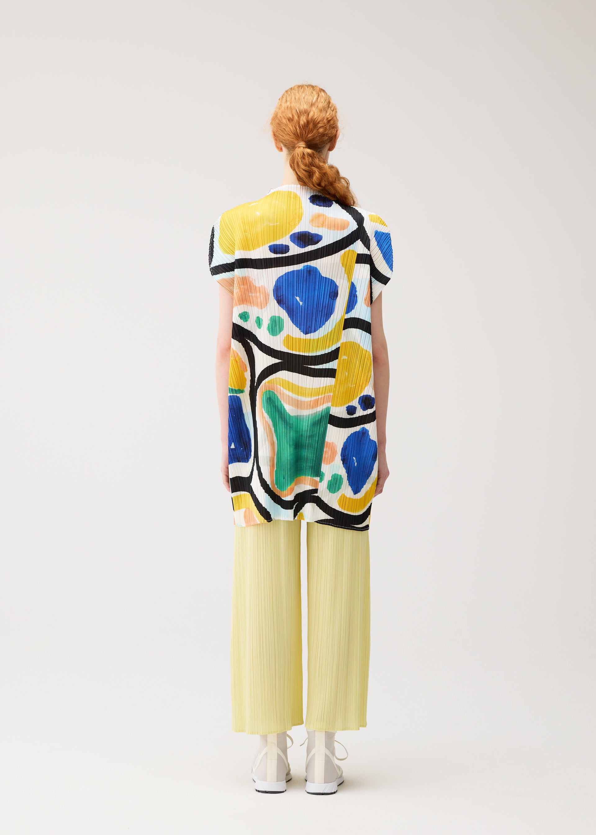 Zoomed view A person with long red hair in a braid wears the ROLL tunic by PLEATS PLEASE ISSEY MIYAKE, paired with light yellow pleated pants and white shoes, standing facing away against a plain light background.