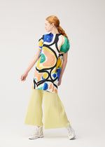 A person with long red hair in a ponytail models the colorful ROLL tunic by PLEATS PLEASE ISSEY MIYAKE, paired with pale yellow pleated wide-leg pants and white sneakers, posing against a plain light background.