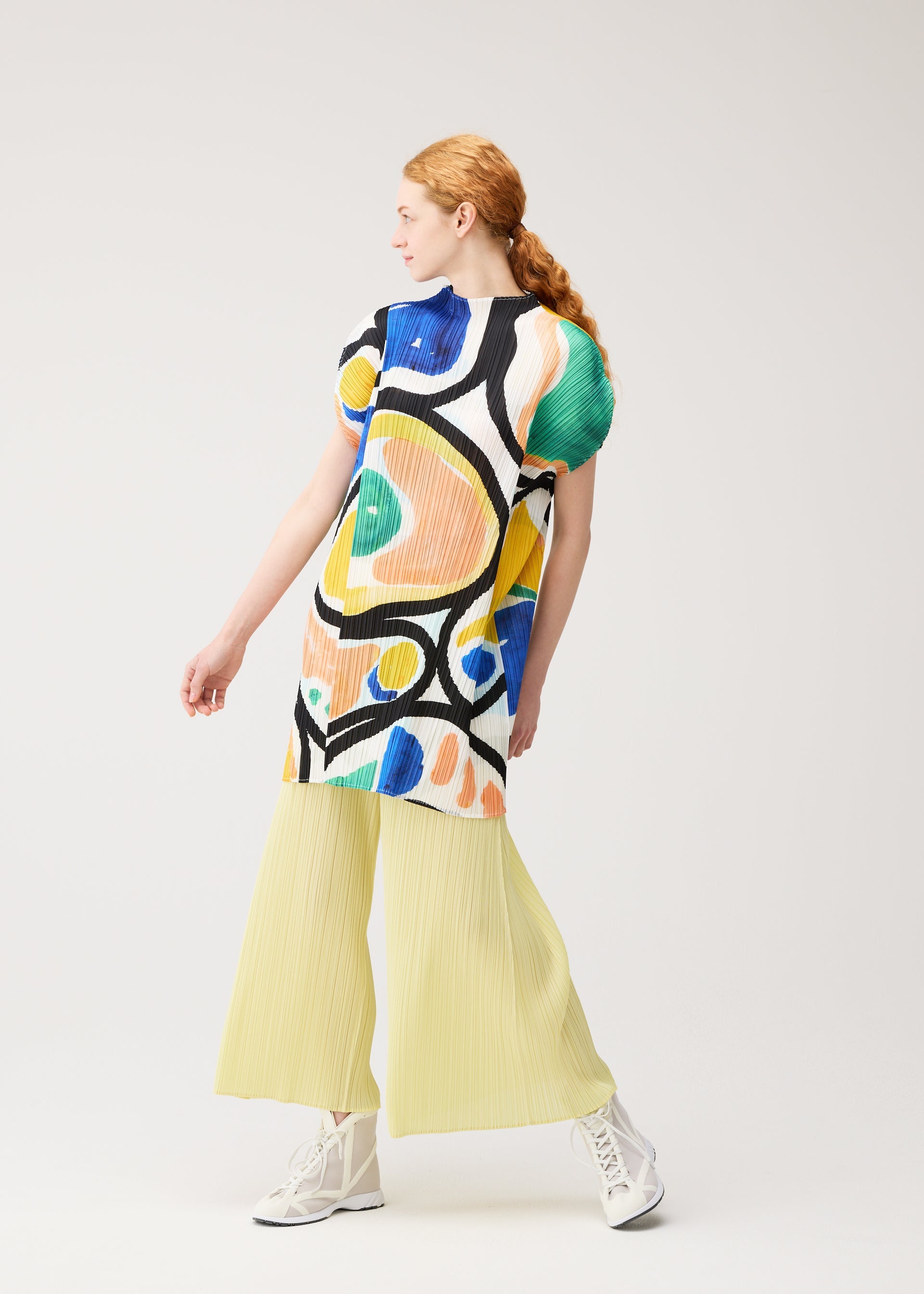 Zoomed view A person with long red hair in a ponytail models the colorful ROLL tunic by PLEATS PLEASE ISSEY MIYAKE, paired with pale yellow pleated wide-leg pants and white sneakers, posing against a plain light background.