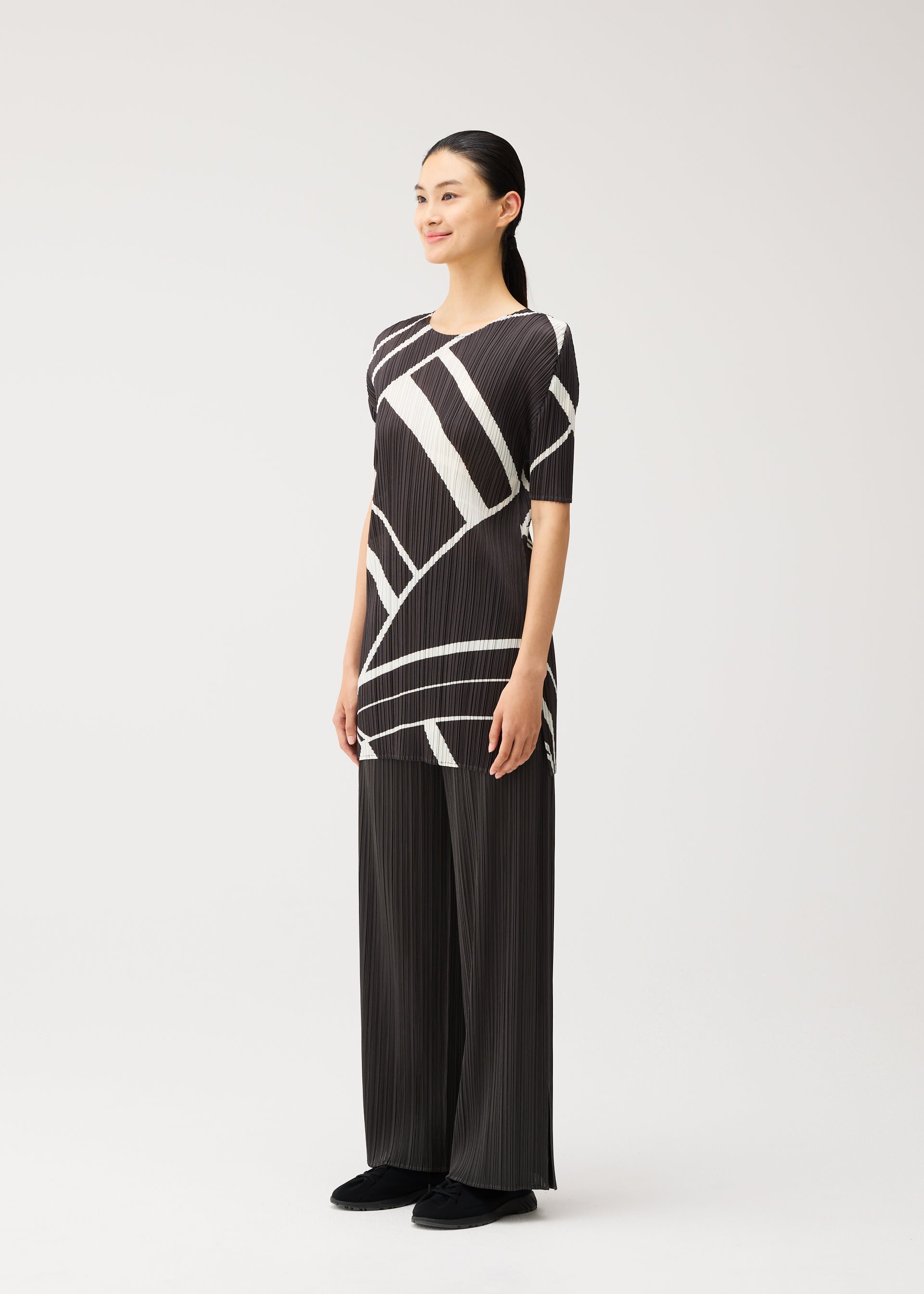 Zoomed view In a minimalist studio, a person stands facing slightly to the left and smiling, dressed in the PLEATS PLEASE ISSEY MIYAKE FOLD LINE collection: a black and white geometric-patterned short-sleeved tunic paired with wide black pleated pants and black shoes.