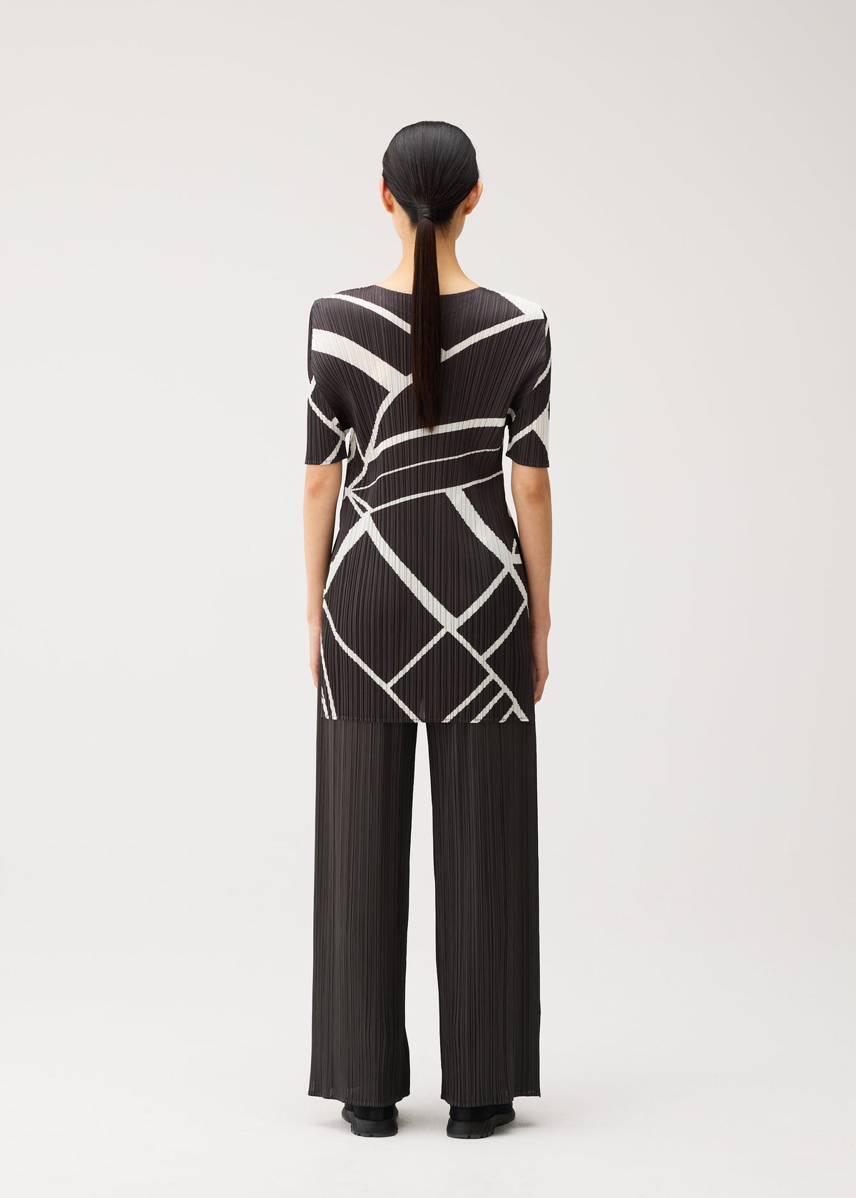 A person with long dark hair in a low ponytail, wearing the FOLD LINE round-neck tunic and wide-leg pleated pants by PLEATS PLEASE ISSEY MIYAKE, featuring a black and white geometric pattern, stands facing away against a plain light background. view 7 of 8