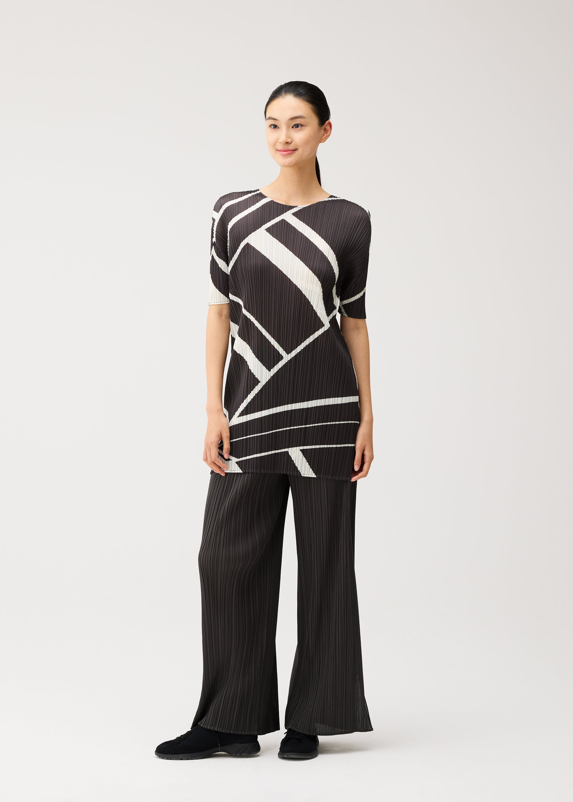 Zoomed view Against a plain background, a person wears the FOLD LINE tunic by PLEATS PLEASE ISSEY MIYAKE—featuring a black and white geometric pattern, round neckline, and elbow-length sleeves—paired with loose black pants. Their dark hair is tied back and they smile slightly, arms relaxed at their sides. The overall look reflects a minimalist aesthetic.