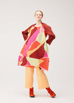 A person dressed in the PLEATS PLEASE ISSEY MIYAKE SURPRISE! collection, featuring a colorful, abstract-patterned pleated tunic with red, pink, and green tones, orange pleated pants, red boots, and a red jacket draped over their shoulders, stands against a plain background.