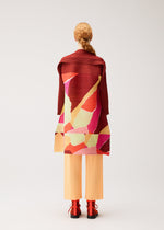 A person with long, light hair in a low ponytail stands facing away from the camera against a plain white background, wearing the PLEATS PLEASE ISSEY MIYAKE SURPRISE! colorful geometric-patterned tunic, peach pleated pants, and red shoes.