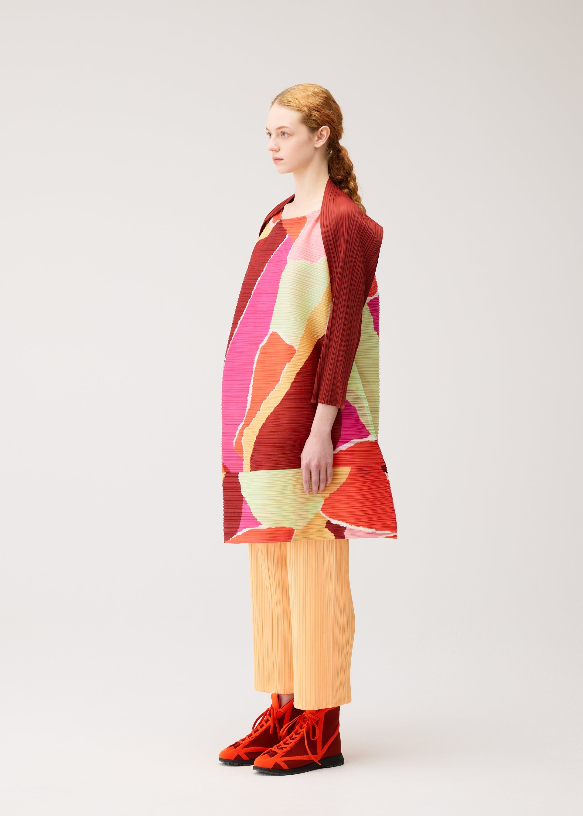 Zoomed view A woman stands in profile against a plain light background, wearing the PLEATS PLEASE ISSEY MIYAKE “SURPRISE!” colorful boat-neck tunic featuring abstract red, pink, green, and yellow patterns, paired with light orange pleated pants and red shoes.