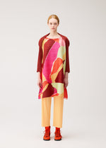 A person stands facing forward against a plain light background, wearing “SURPRISE!” by PLEATS PLEASE ISSEY MIYAKE—a colorful, abstract-patterned boat-neck tunic styled as a dress—paired with orange pleated pants, a red jacket draped over their shoulders, and bright orange shoes.