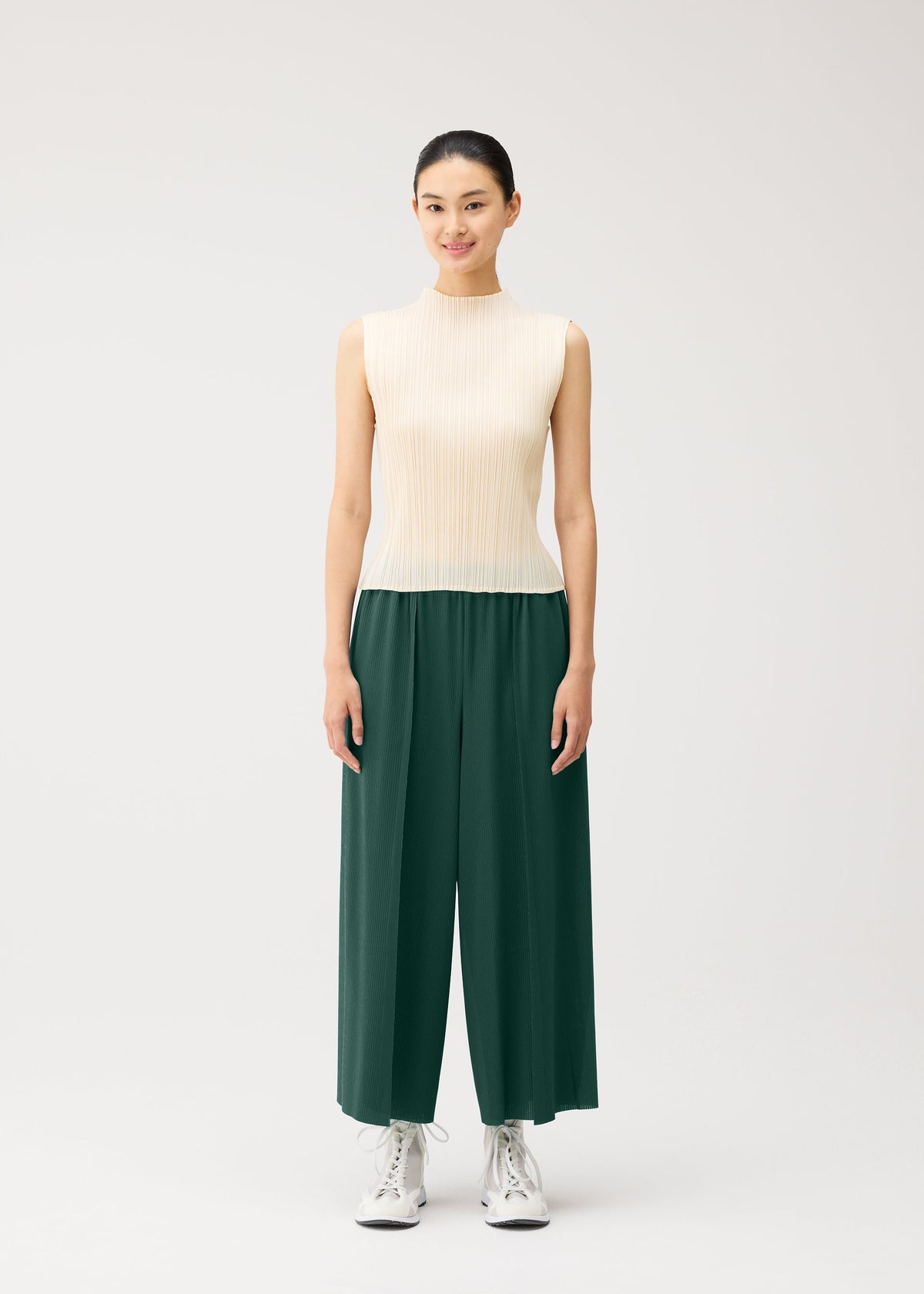 A woman stands facing forward, wearing a sleeveless beige top, white sneakers, and the PLEATS PLEASE ISSEY MIYAKE A-POC BOTTOMS—wide-leg dark green pleated pants—against a plain light background. view 9 of 13