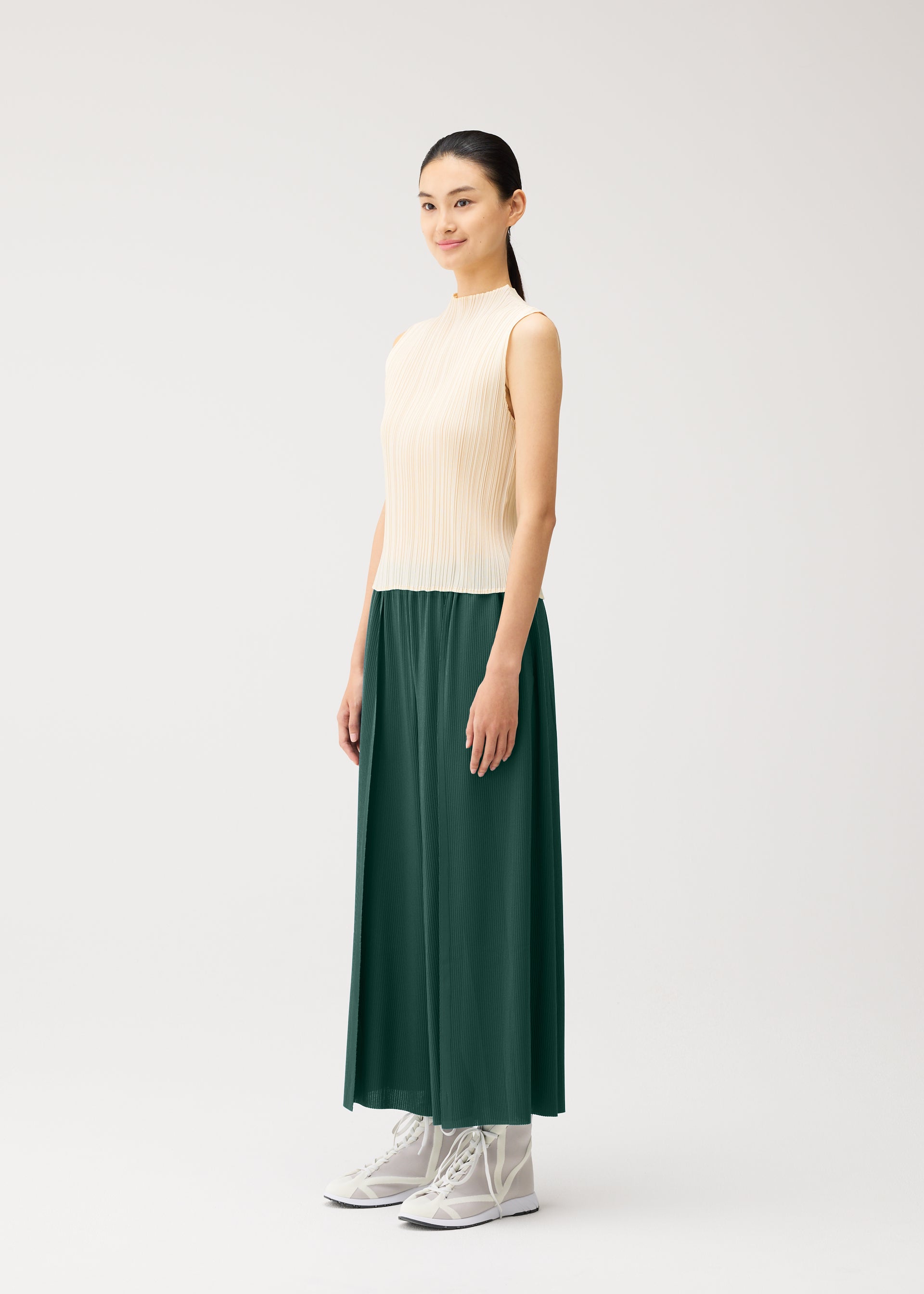 Zoomed view A woman stands in a neutral studio, wearing the PLEATS PLEASE ISSEY MIYAKE A-POC BOTTOMS—a long green skirt with a gentle flared and tapered silhouette—paired with a sleeveless beige top and light gray high-top shoes. She has straight dark hair tied back and is smiling slightly.