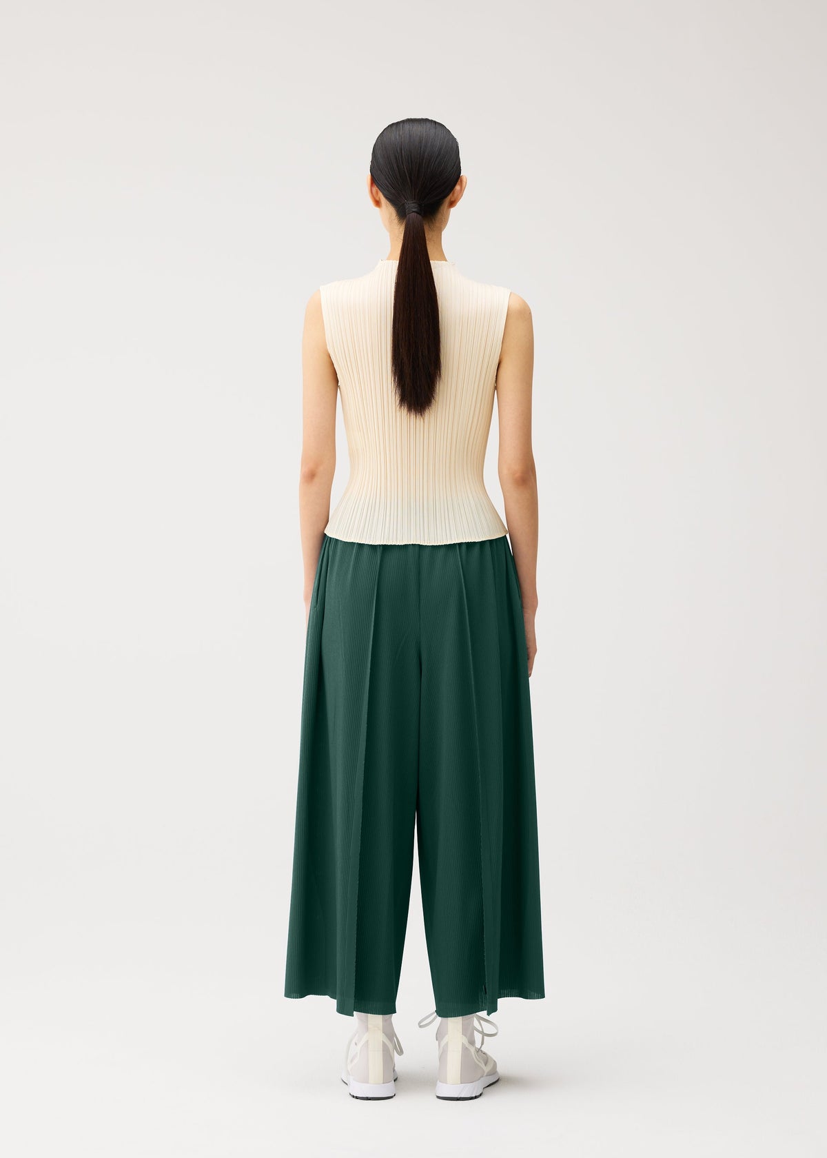 A person with long dark hair in a ponytail stands facing away, wearing a sleeveless beige ribbed top, white shoes, and PLEATS PLEASE ISSEY MIYAKE A-POC BOTTOMS—flared and tapered green pleated pants—against a plain light background. view 11 of 13