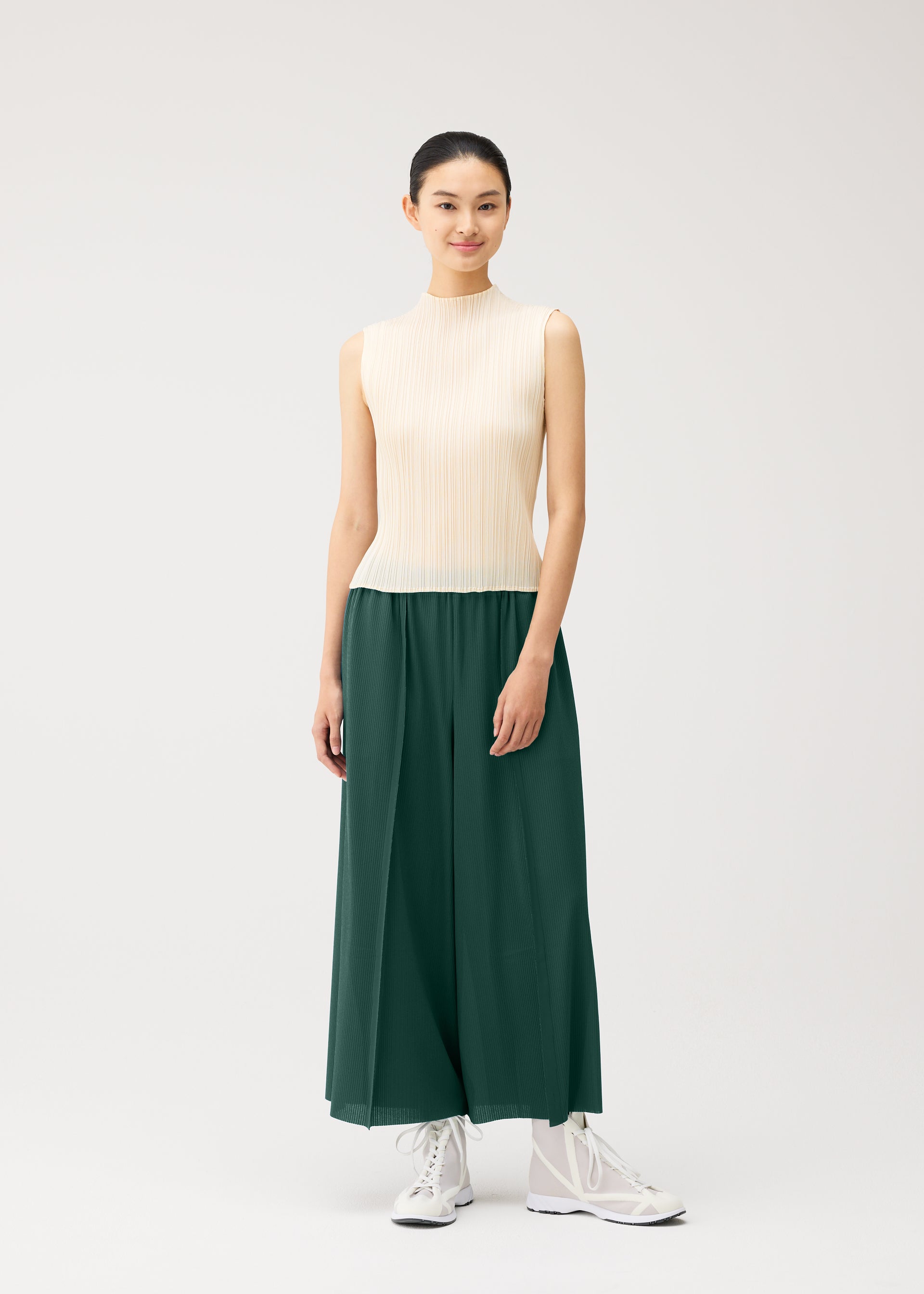 Zoomed view A woman with dark hair tied back and a gentle smile stands against a light background, wearing wide-leg green A-POC BOTTOMS from PLEATS PLEASE ISSEY MIYAKE, paired with a sleeveless cream top and white sneakers.