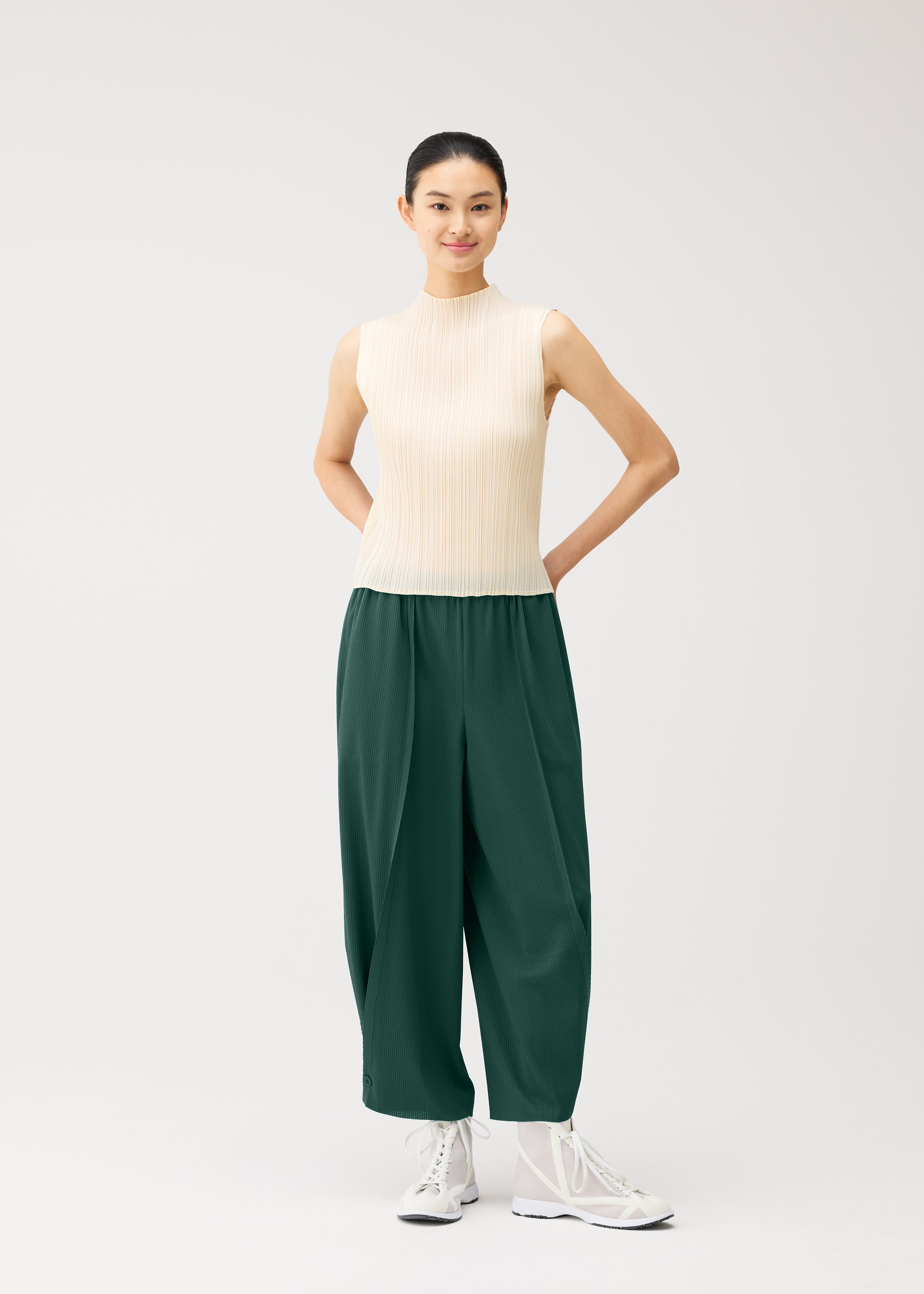 Zoomed view Wearing the PLEATS PLEASE ISSEY MIYAKE A-POC BOTTOMS, a person in a sleeveless beige top and white sneakers stands against a plain light background, smiling with their hands behind their back.