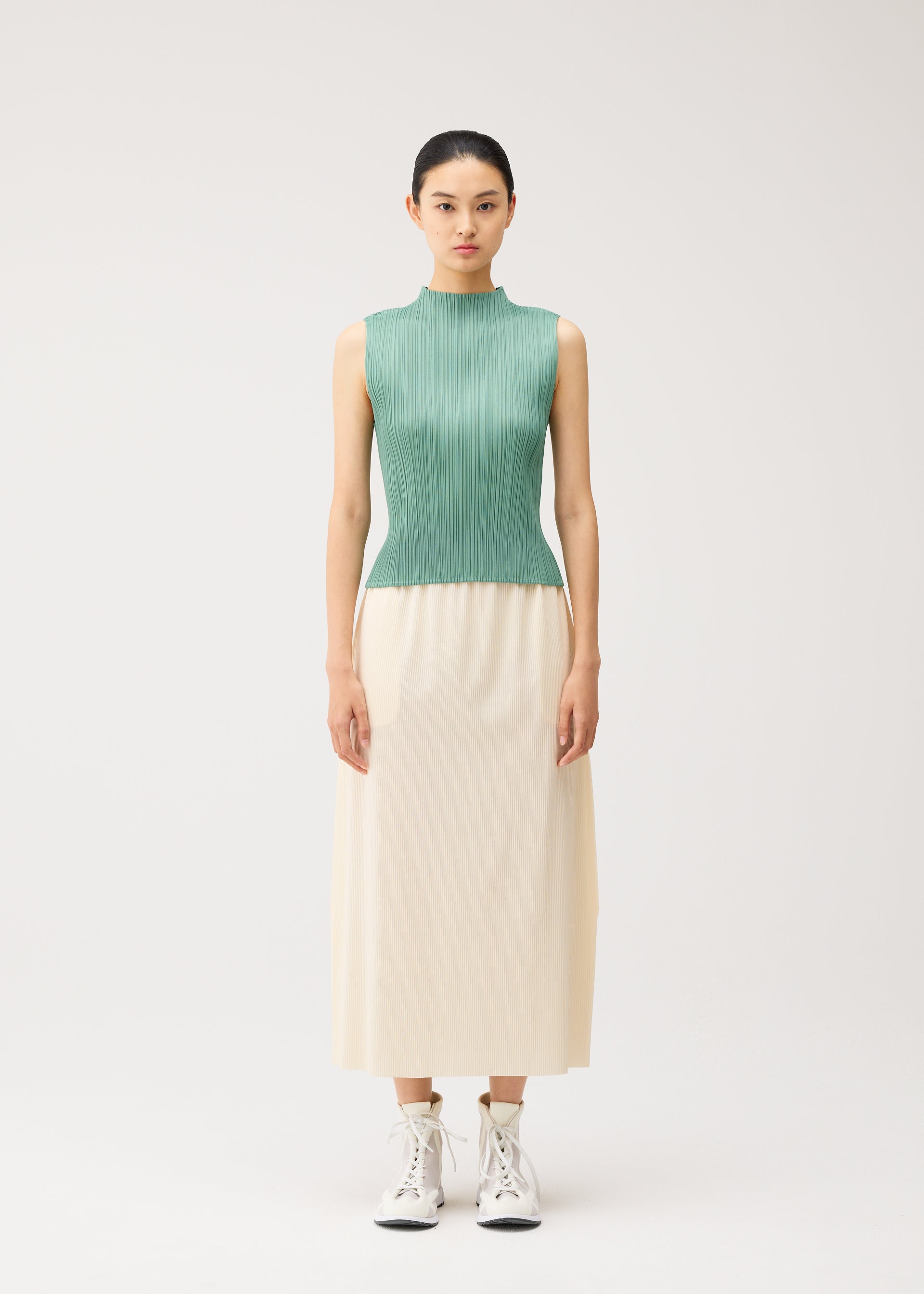 Zoomed view A woman stands facing forward, wearing a sleeveless green pleated top and PLEATS PLEASE ISSEY MIYAKE A-POC BOTTOMS—a long cream skirt with pockets—paired with white lace-up boots, against a plain light background.