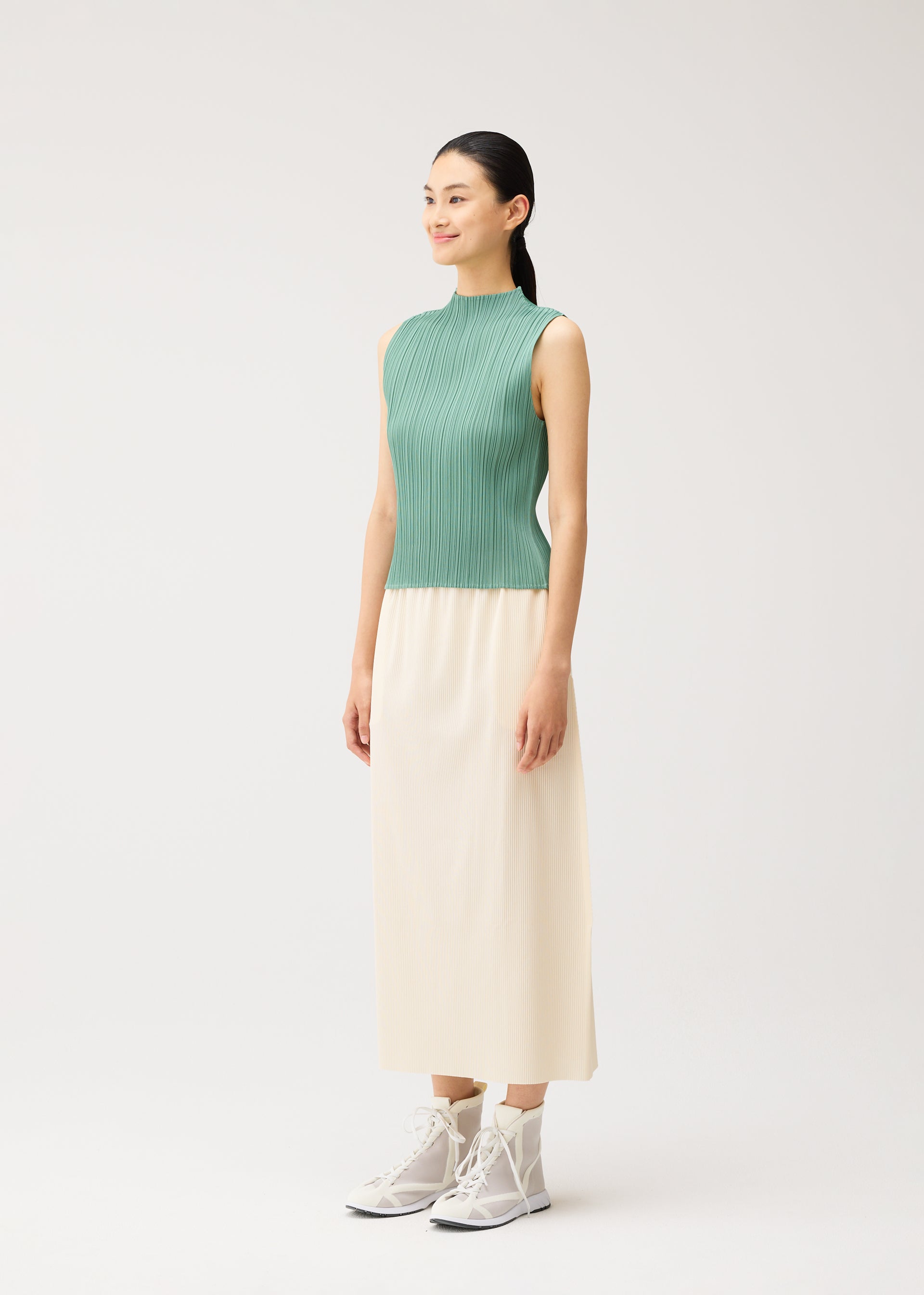 Zoomed view A woman stands against a plain light background, wearing PLEATS PLEASE ISSEY MIYAKE’s A-POC BOTTOMS—a cream pleated skirt—paired with a green sleeveless top and white high-top sneakers.