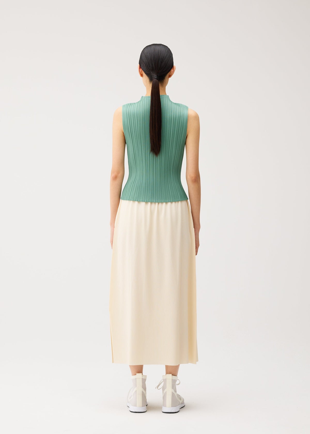 A person with long dark hair in a ponytail, dressed in a sleeveless green ribbed top and the A-POC BOTTOMS by PLEATS PLEASE ISSEY MIYAKE—a long cream pleated skirt—stands facing away in a plain, light studio. view 8 of 9