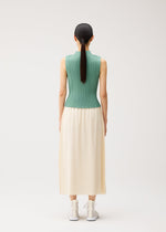 A person with long dark hair in a ponytail, dressed in a sleeveless green ribbed top and the A-POC BOTTOMS by PLEATS PLEASE ISSEY MIYAKE—a long cream pleated skirt—stands facing away in a plain, light studio.
