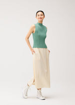 A woman stands smiling against a plain light background, wearing a sleeveless green pleated top, PLEATS PLEASE ISSEY MIYAKE A-POC BOTTOMS—a long cream pleated skirt with pockets—and white high-top sneakers.