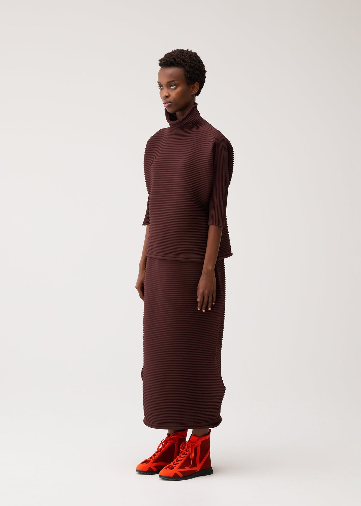 A person stands in profile against a plain light background, dressed in the ZIG ZAG KNIT set from PLEATS PLEASE ISSEY MIYAKE—a ribbed burgundy turtleneck top and matching seamless skirt with a subtle zigzag silhouette—paired with bright red high-top sneakers. view 6 of 8