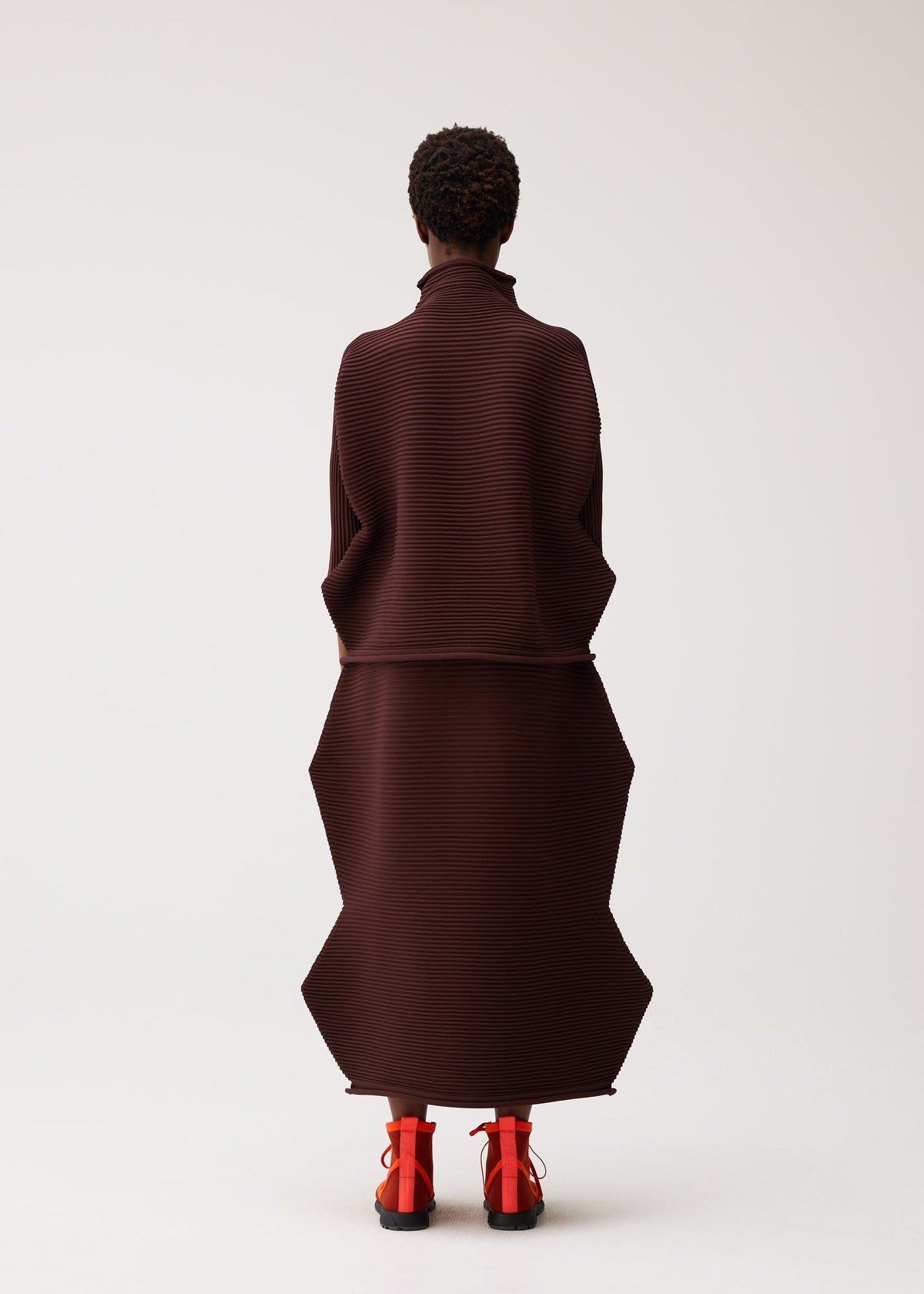 A person stands facing away, wearing the PLEATS PLEASE ISSEY MIYAKE ZIG ZAG KNIT—a structured, dark brown pleated outfit featuring geometric shapes and a zigzag silhouette—paired with bright red shoes, against a plain light background. view 7 of 8