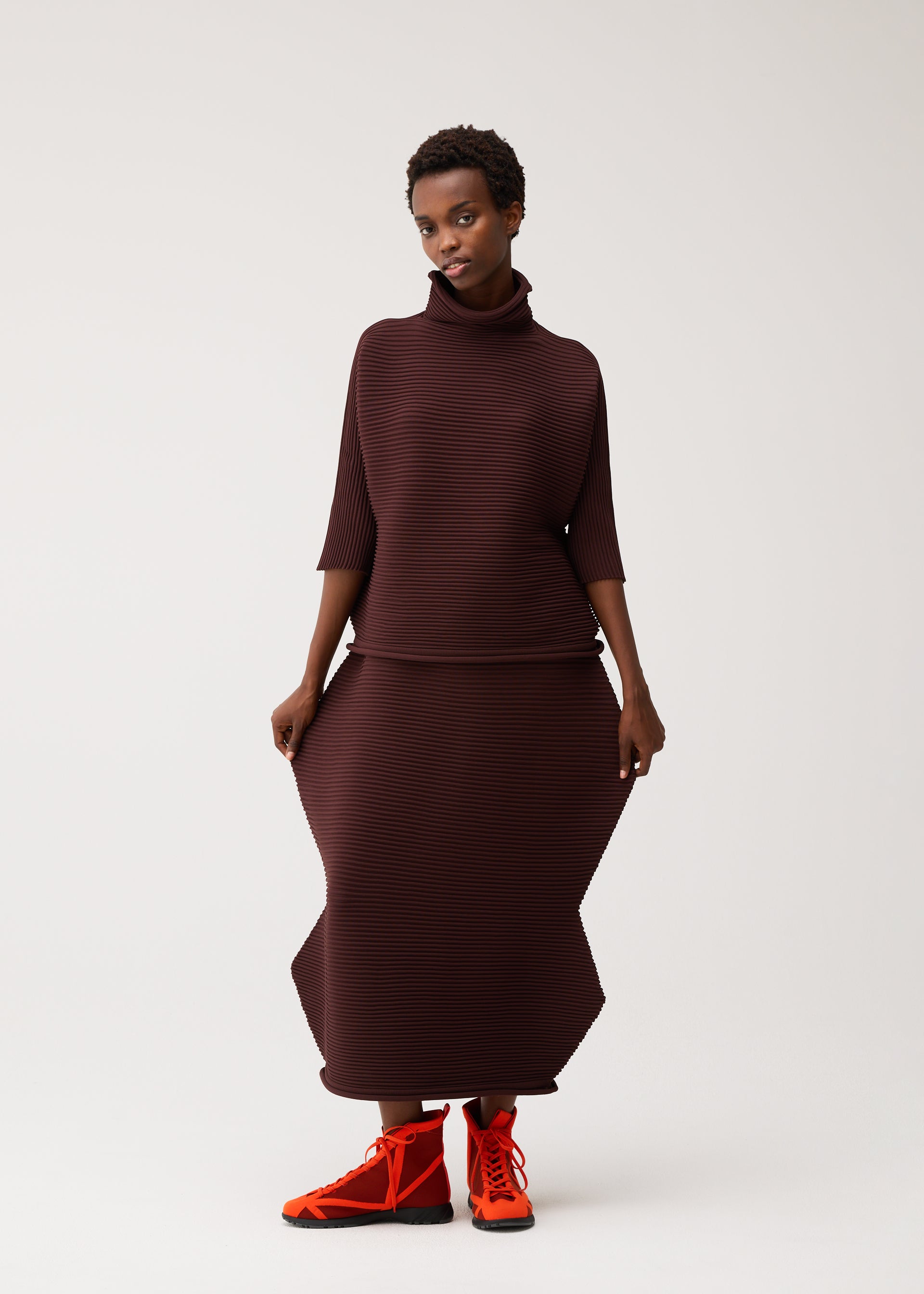 Zoomed view Wearing the ZIG ZAG KNIT two-piece outfit by PLEATS PLEASE ISSEY MIYAKE, a person stands against a plain white background. The ensemble features a brown, textured high-neck top and a long, sculptural skirt crafted in a seamless knit design that creates a striking zigzag silhouette. Bright orange lace-up shoes complete the bold look.