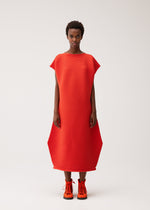 A person stands against a plain background, looking directly at the camera, wearing the PLEATS PLEASE ISSEY MIYAKE ZIG ZAG KNIT—a red, textured geometric round-neck dress with a bold zigzag silhouette—paired with bright orange lace-up shoes.