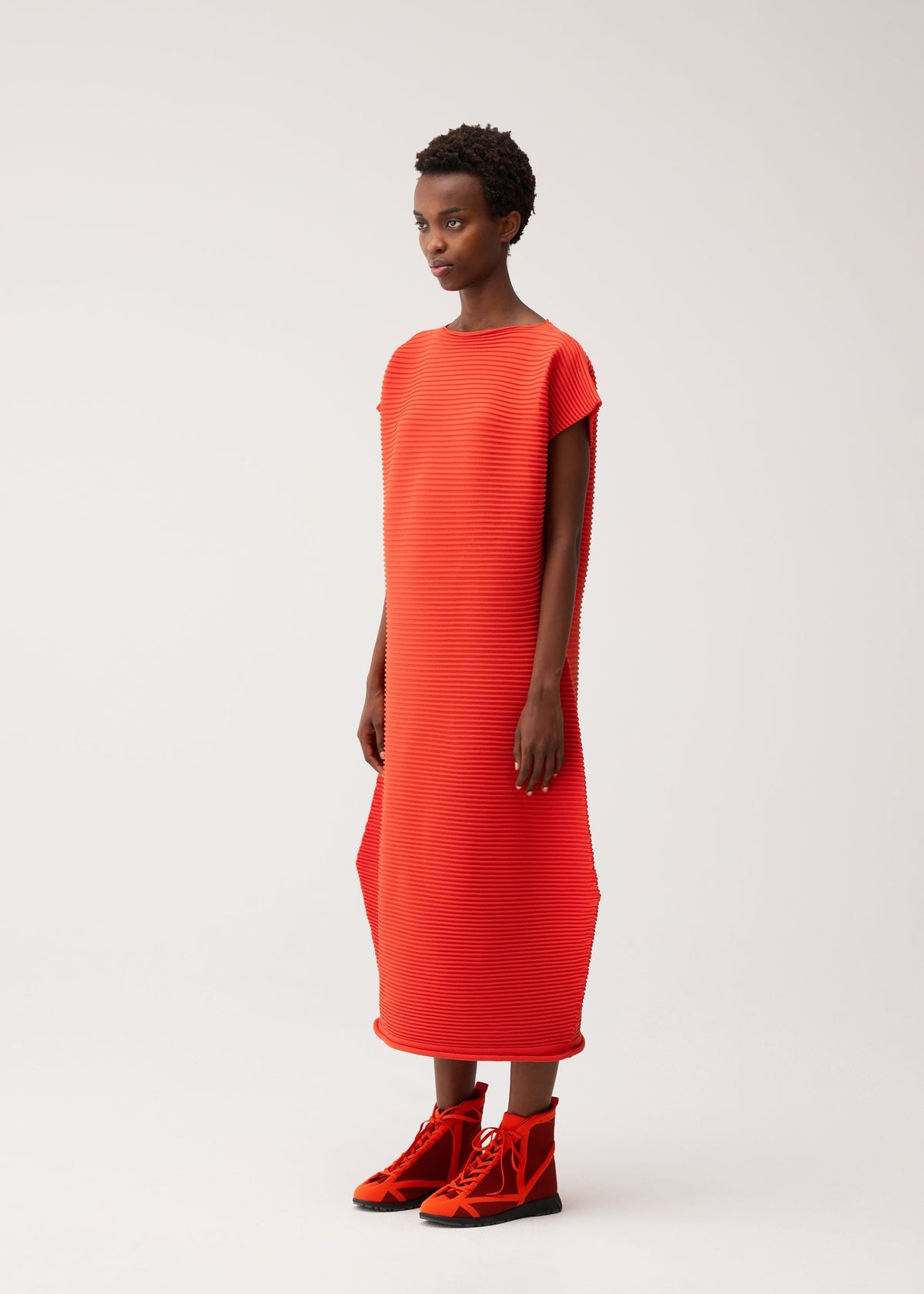 A person is wearing the PLEATS PLEASE ISSEY MIYAKE ZIG ZAG KNIT, a loose, bright orange seamless knit dress with a zigzag silhouette and short sleeves, paired with matching orange high-top shoes. They have short hair and are standing against a plain white background. view 6 of 8