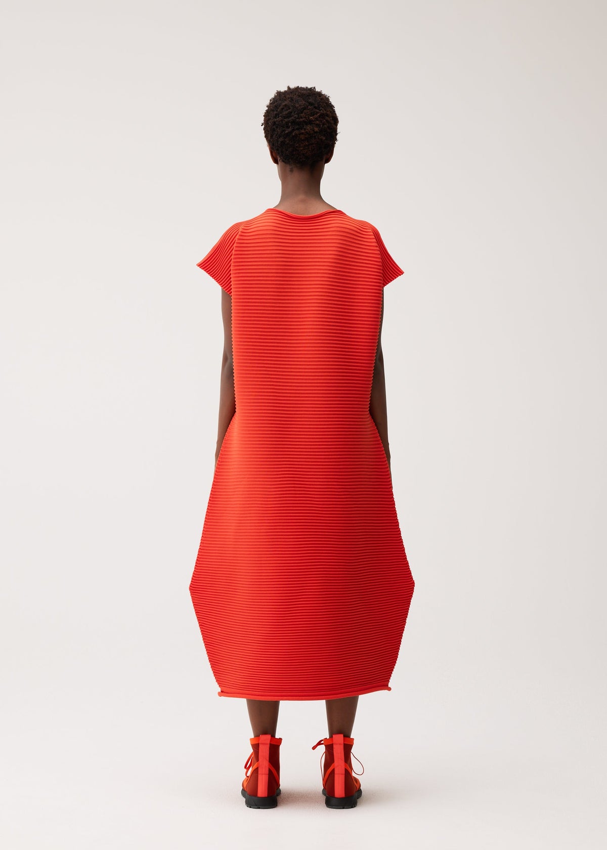 A person stands with their back to the camera, wearing the PLEATS PLEASE ISSEY MIYAKE ZIG ZAG KNIT dress—a textured, seamless bright red design with a round neckline—paired with matching red shoes, set against a plain light background. view 7 of 8