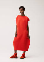 A woman stands smiling against a plain white background, wearing the PLEATS PLEASE ISSEY MIYAKE ZIG ZAG KNIT—a bright red, textured, loose-fitting round-neck dress with a distinctive zigzag silhouette—paired with matching red high-top sneakers.