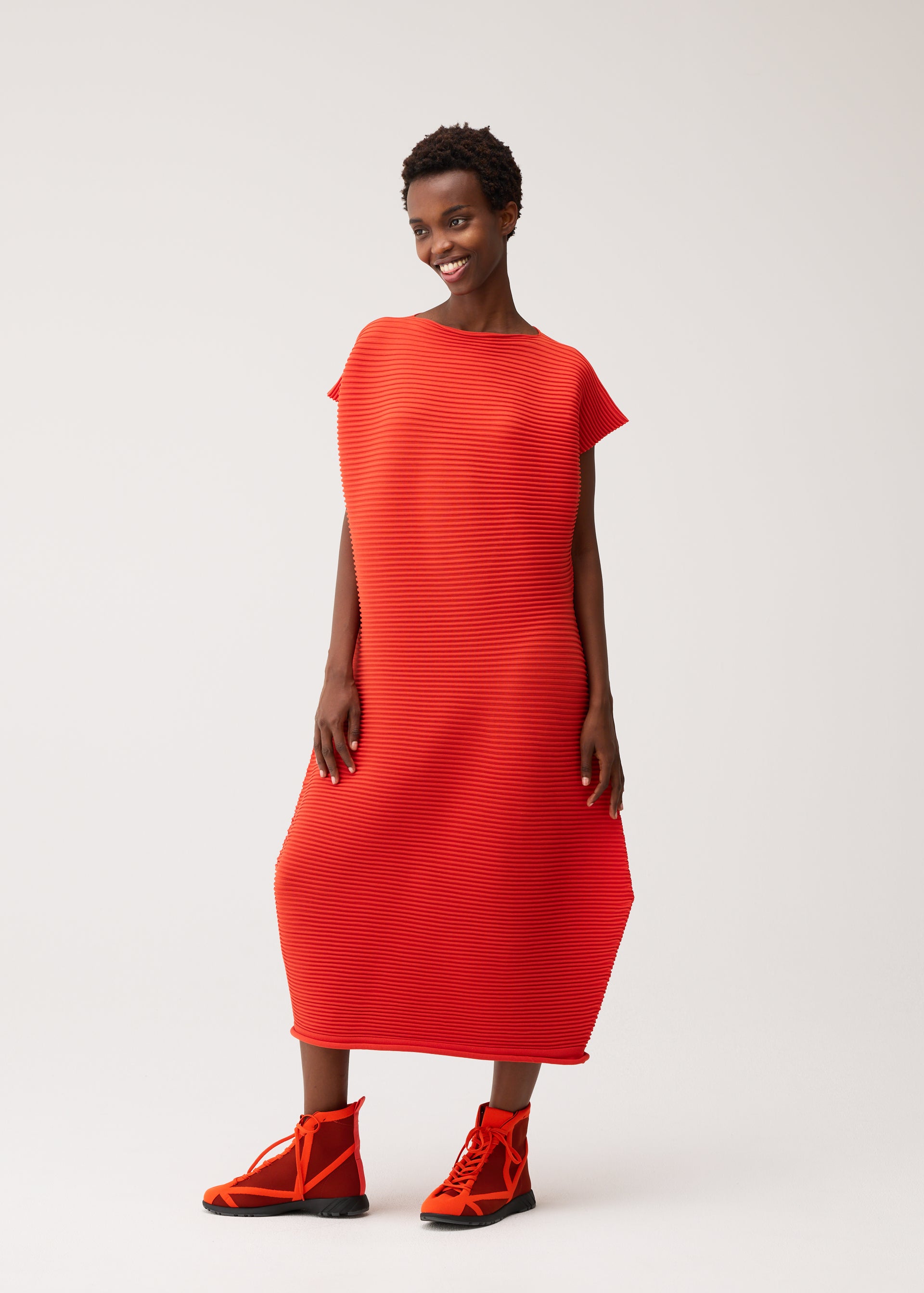 Zoomed view A woman stands smiling against a plain white background, wearing the PLEATS PLEASE ISSEY MIYAKE ZIG ZAG KNIT—a bright red, textured, loose-fitting round-neck dress with a distinctive zigzag silhouette—paired with matching red high-top sneakers.