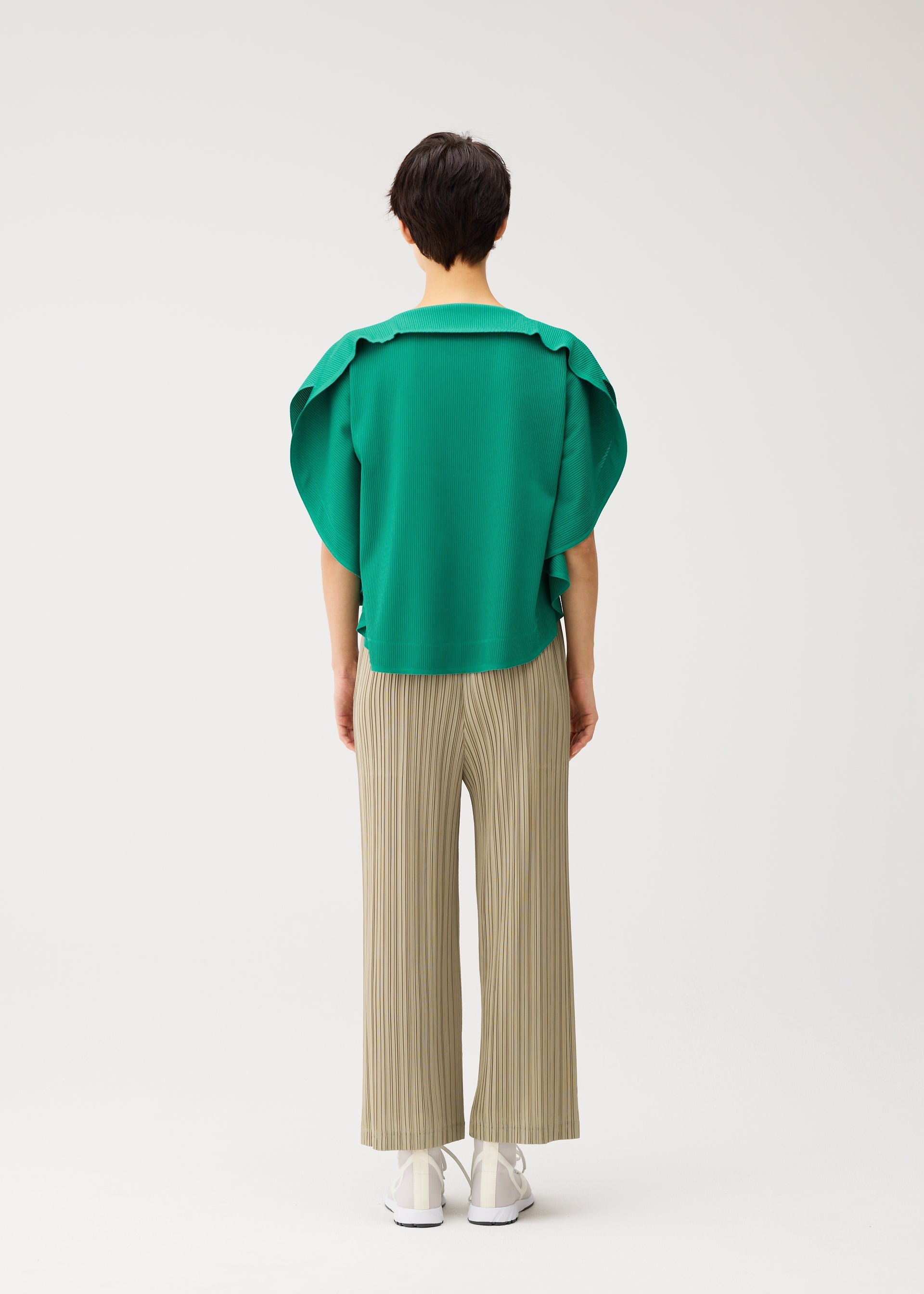 Zoomed view A person with short dark hair faces away, wearing a loose, round-neck A-POC SLICE top with wide sleeves from PLEATS PLEASE ISSEY MIYAKE, light beige pleated pants, and white shoes against a plain white background.