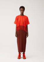 A person stands against a plain background, wearing the PLEATS PLEASE ISSEY MIYAKE A-POC LACE—a loose red fringed top featuring a subtle striped pattern at the hem—paired with a brown pleated skirt and bright orange lace-up shoes. They have short curly hair and a neutral expression.