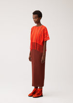 A woman stands against a plain light background wearing the PLEATS PLEASE ISSEY MIYAKE A-POC LACE ensemble, featuring a fringed top, pleated skirt, and red shoes.