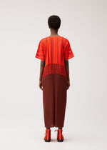 A person stands facing away, wearing the PLEATS PLEASE ISSEY MIYAKE A-POC LACE top in a red and orange striped pattern with fringed details, paired with a brown pleated skirt. They have short curly hair and red sandals, set against a plain, light-colored background.