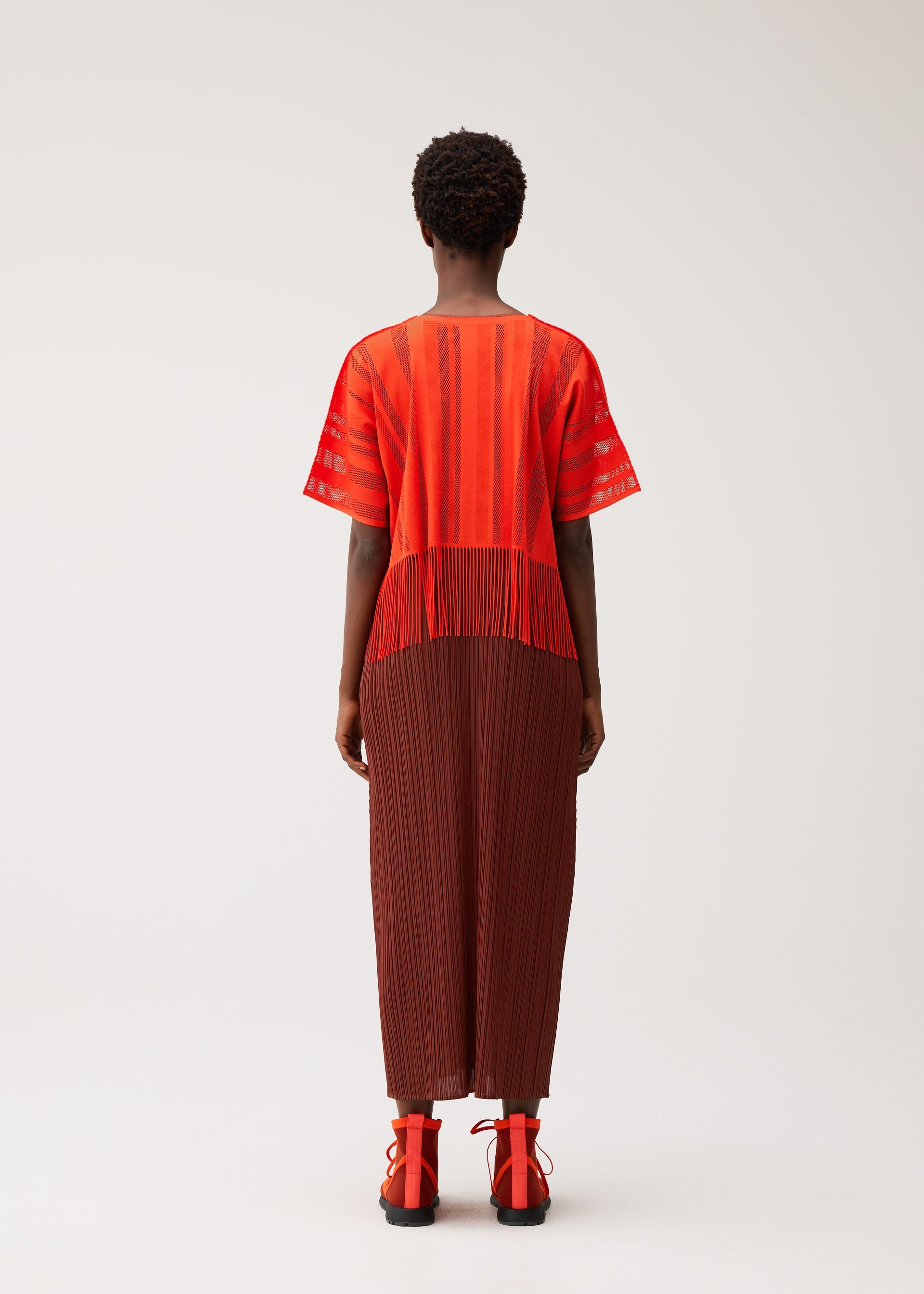 Zoomed view A person stands facing away, wearing the PLEATS PLEASE ISSEY MIYAKE A-POC LACE top in a red and orange striped pattern with fringed details, paired with a brown pleated skirt. They have short curly hair and red sandals, set against a plain, light-colored background.