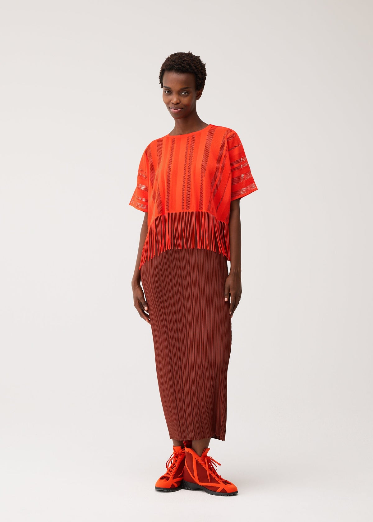 A woman stands against a plain light background wearing the PLEATS PLEASE ISSEY MIYAKE A-POC LACE red fringed short-sleeve top with a subtle striped pattern, paired with a long brown pleated skirt and bright orange shoes. view 7 of 8