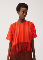 A person wears the PLEATS PLEASE ISSEY MIYAKE A-POC LACE bright orange mesh top, featuring sheer striped panels and fringe at the hem, paired with a dark brown pleated skirt, standing against a plain light background.