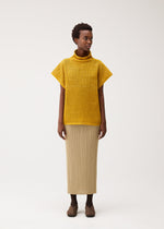 A person stands against a plain light background wearing the yellow DOT KNIT by PLEATS PLEASE ISSEY MIYAKE, paired with a beige pleated skirt and brown shoes.