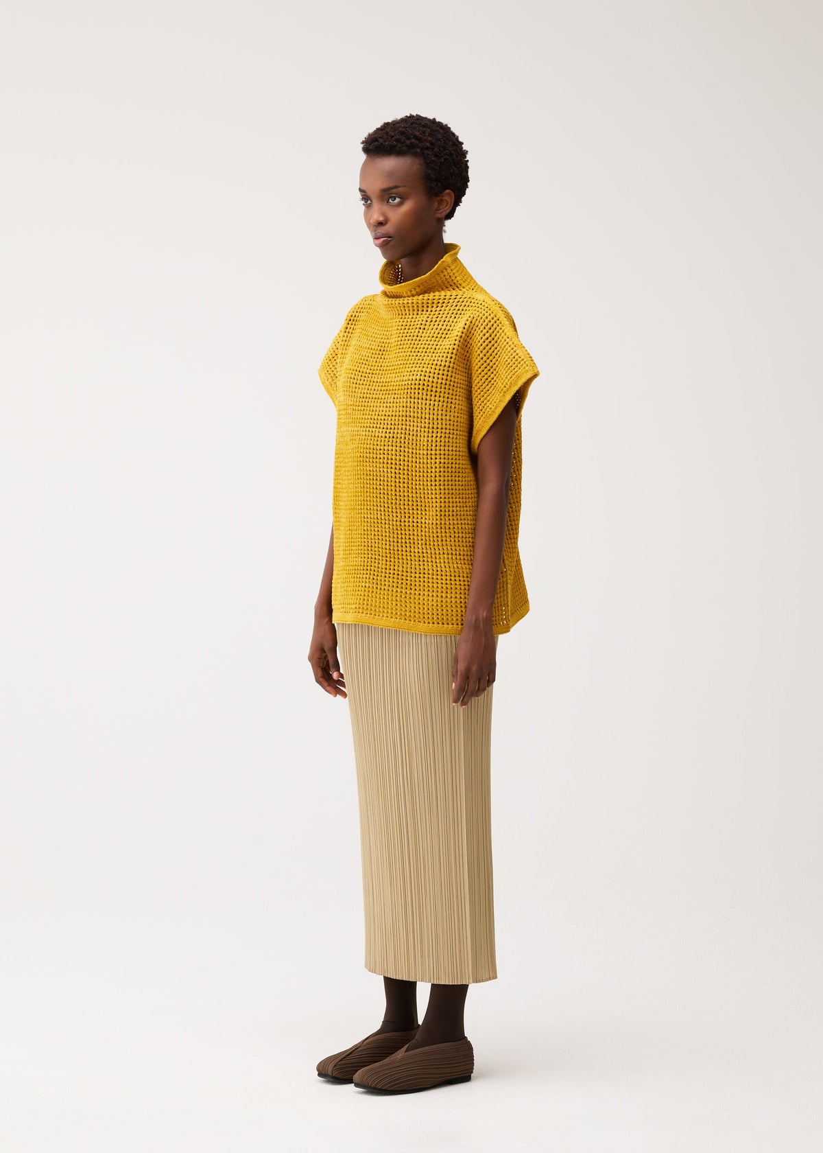 Zoomed view A woman faces left against a white background, wearing the DOT KNIT by PLEATS PLEASE ISSEY MIYAKE, paired with a beige pleated skirt, dark tights, and brown slippers.