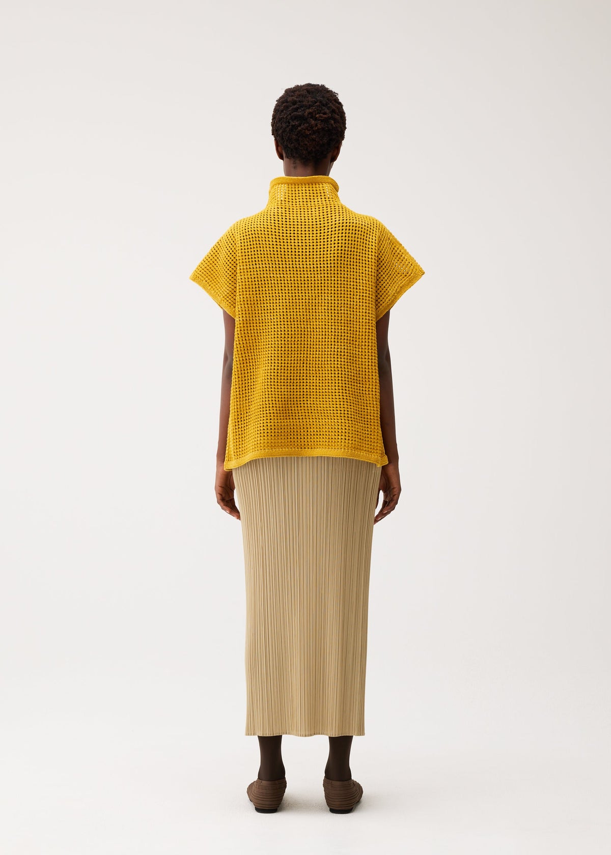 A person stands facing away, wearing the yellow DOT KNIT by PLEATS PLEASE ISSEY MIYAKE and a beige ankle-length pleated skirt with brown shoes, against a plain light background. view 7 of 8