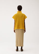 A person stands facing away, wearing the yellow DOT KNIT by PLEATS PLEASE ISSEY MIYAKE and a beige ankle-length pleated skirt with brown shoes, against a plain light background.