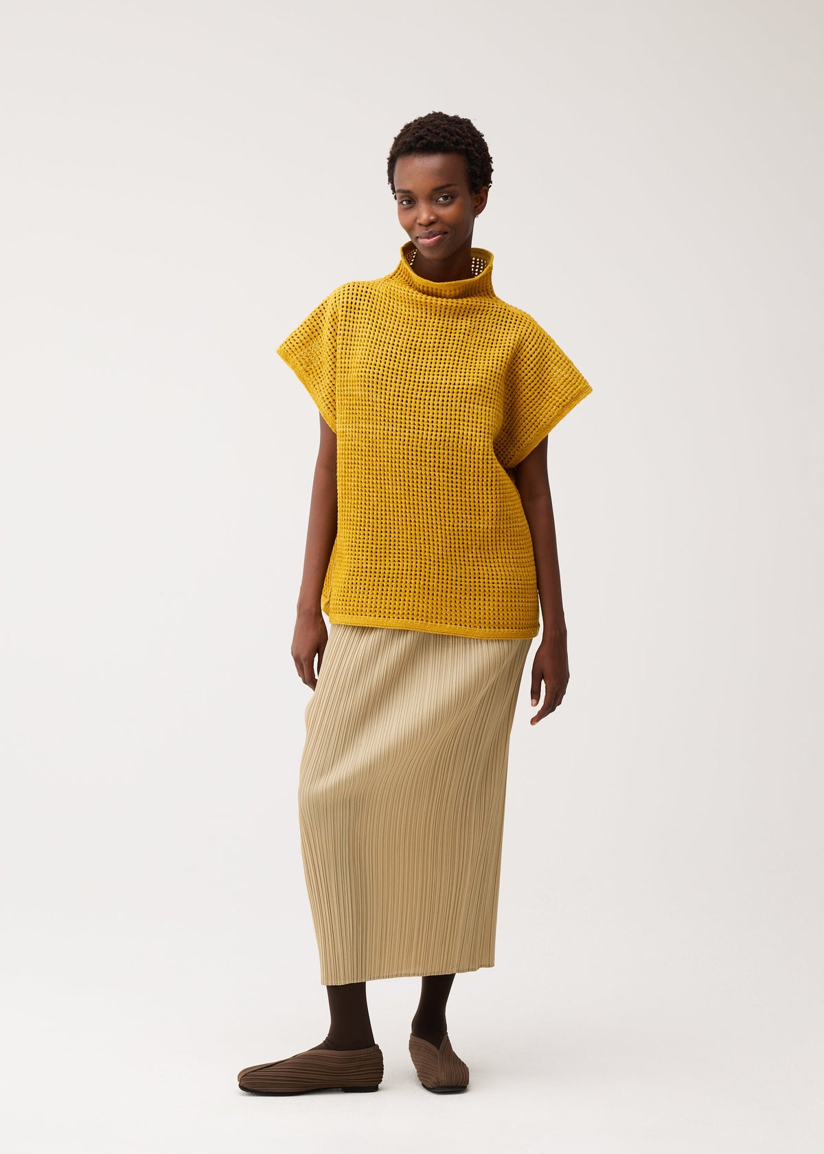 Wearing the DOT KNIT by PLEATS PLEASE ISSEY MIYAKE—a yellow textured high-neck short-sleeve top—this person pairs it with a beige pleated skirt, dark socks, and brown shoes, smiling softly against a plain background. view 8 of 8