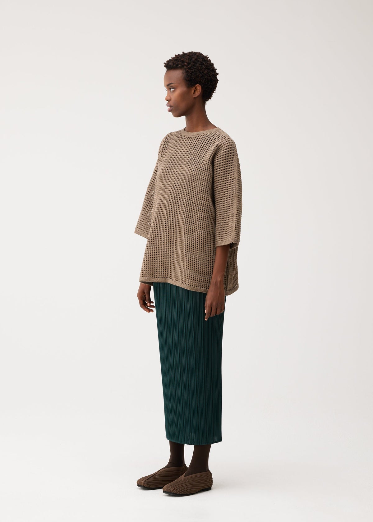 A person stands sideways, wearing the DOT KNIT by PLEATS PLEASE ISSEY MIYAKE, a loose beige round-neck mesh-patterned knit, styled with a green pleated midi skirt and brown shoes against a plain light background. view 6 of 8