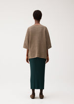 A person faces away, wearing the DOT KNIT by PLEATS PLEASE ISSEY MIYAKE—a loose, round-neck brown knit with a mesh-like pattern—paired with a long pleated dark green skirt and brown shoes.