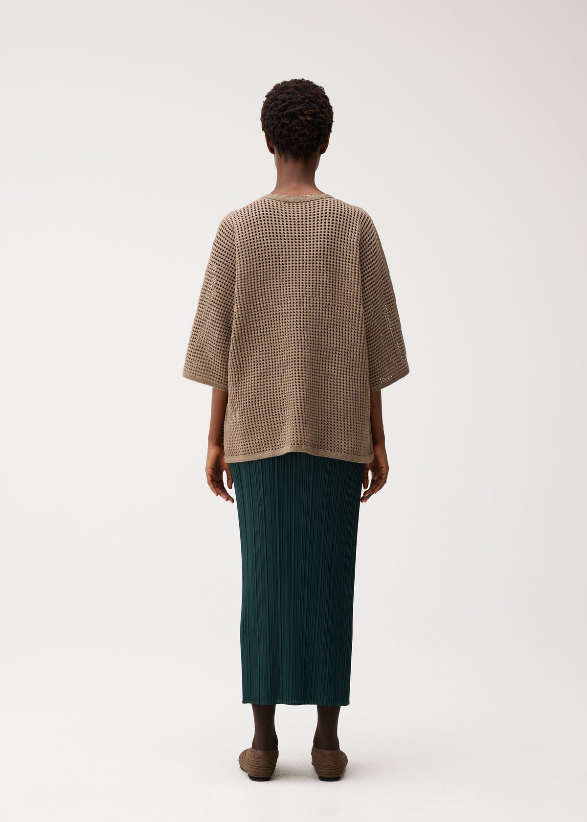 Zoomed view A person faces away, wearing the DOT KNIT by PLEATS PLEASE ISSEY MIYAKE—a loose, round-neck brown knit with a mesh-like pattern—paired with a long pleated dark green skirt and brown shoes.