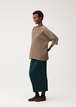 A woman stands sideways, smiling, in the DOT KNIT by PLEATS PLEASE ISSEY MIYAKE—a loose beige round-neck sweater with a mesh pattern—paired with a long dark green pleated skirt and brown shoes.