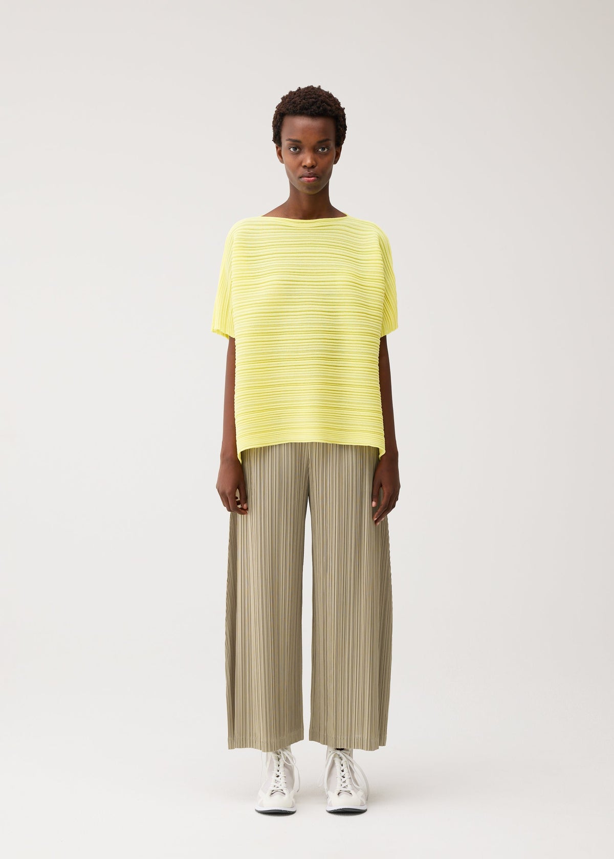 A person stands facing forward against a plain background, wearing the PAPER BOUNCE KNIT wide-legged beige pleated pants from PLEATS PLEASE ISSEY MIYAKE, a loose yellow striped boat-neck top, and white sneakers. view 5 of 8
