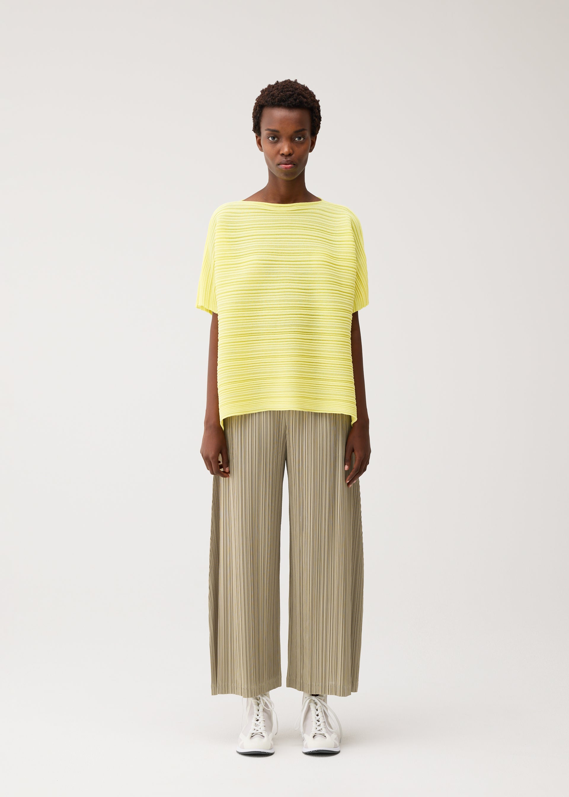 Zoomed view A person stands facing forward against a plain background, wearing the PAPER BOUNCE KNIT wide-legged beige pleated pants from PLEATS PLEASE ISSEY MIYAKE, a loose yellow striped boat-neck top, and white sneakers.