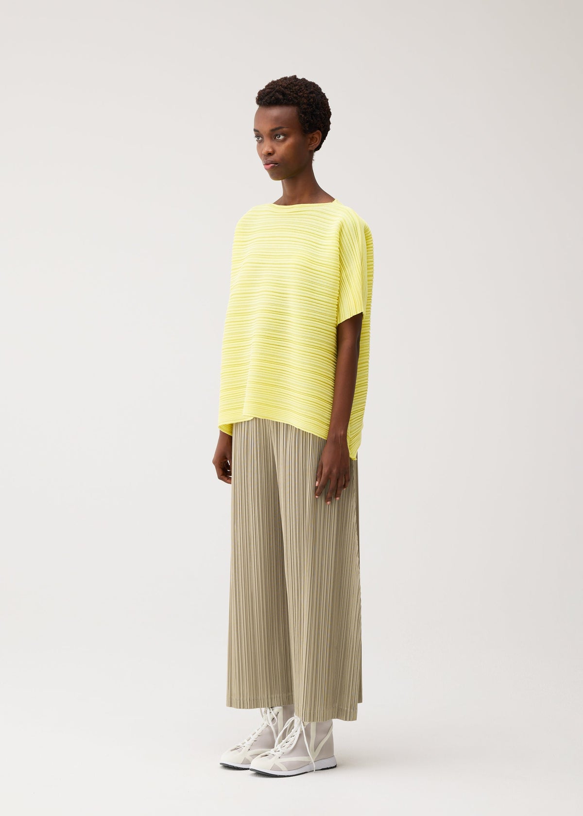 A person with short curly hair and a neutral expression stands against a plain background, wearing the PLEATS PLEASE ISSEY MIYAKE PAPER BOUNCE KNIT yellow boat-neck top, beige pleated wide-leg pants, and white high-top sneakers. view 6 of 8