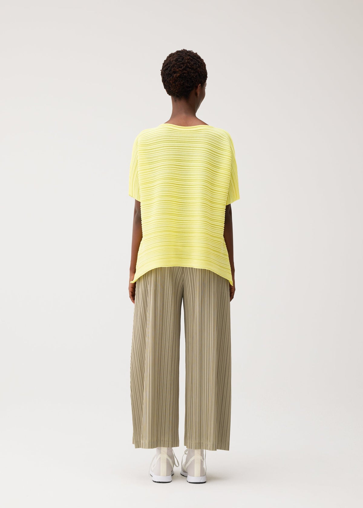 A person with short curly hair faces away, wearing PLEATS PLEASE ISSEY MIYAKE PAPER BOUNCE KNIT—a loose yellow boat-neck top and wide-leg light olive pleated knit pants—with white shoes, set against a plain light background. view 7 of 8