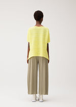 A person with short curly hair faces away, wearing PLEATS PLEASE ISSEY MIYAKE PAPER BOUNCE KNIT—a loose yellow boat-neck top and wide-leg light olive pleated knit pants—with white shoes, set against a plain light background.