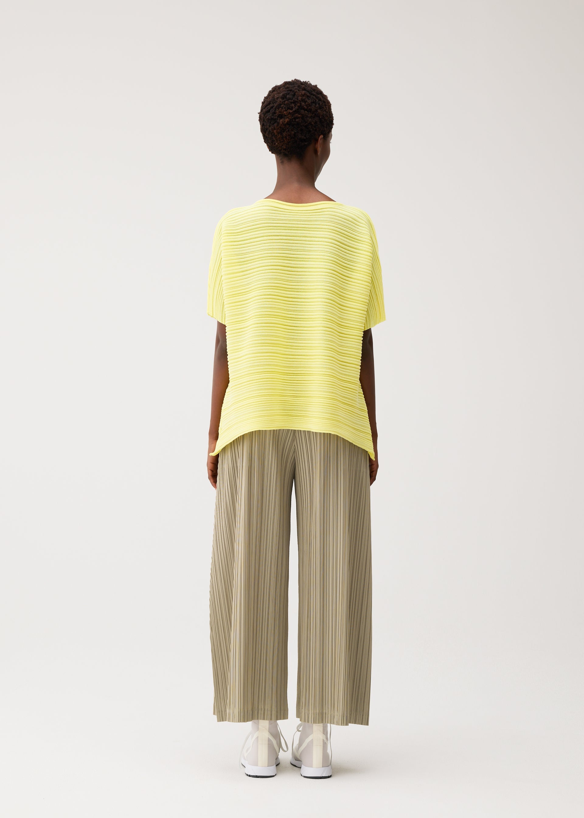 Zoomed view A person with short curly hair faces away, wearing PLEATS PLEASE ISSEY MIYAKE PAPER BOUNCE KNIT—a loose yellow boat-neck top and wide-leg light olive pleated knit pants—with white shoes, set against a plain light background.